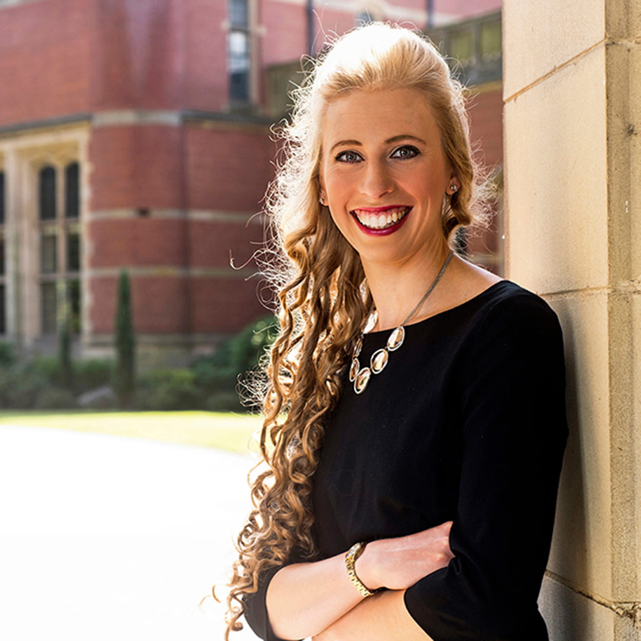 Alice Kinder is a white woman with long, curly blonde hair. She is smiling widely and stands outside, in front of a grand, brick building. She wears a long-sleeved black dress and a silver necklace.