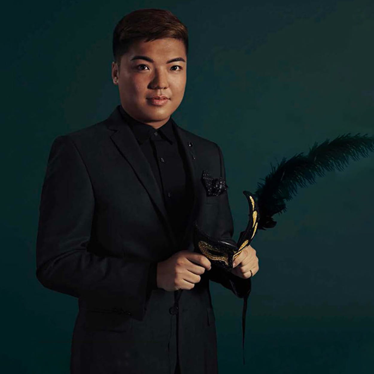 Billy Yu Lok Ng is a man of East Asian descent with short, dark brown hair. He wears a black suit with a black shirt and holds a gold and black feathered theatre mask.