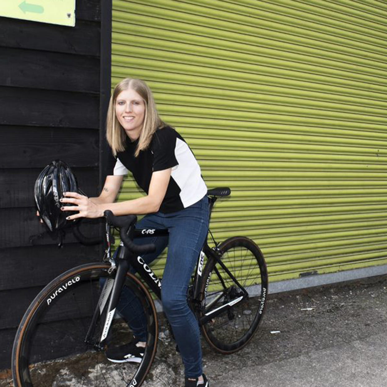 Clare is a blonde white woman perched on a black bicycle wearing a sporty t-shirt and skinny jeans with black and white trainers. She is holding a black bicycle helmet, and sits in front of a chartreuse shutter door.