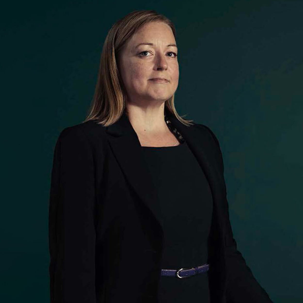 Gemma Lodge was a white woman with long, straight, dark blonde hair. She wears a black dress with a black blazer and leather belt.