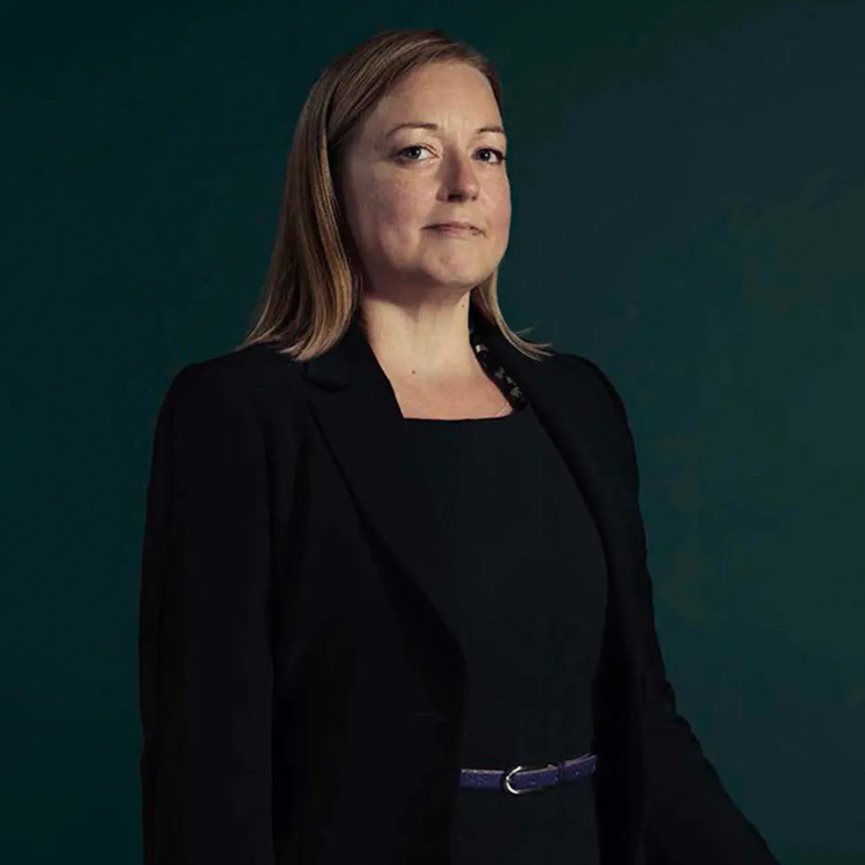 Gemma Lodge was a white woman with long, straight, dark blonde hair. She wears a black dress with a black blazer and leather belt.