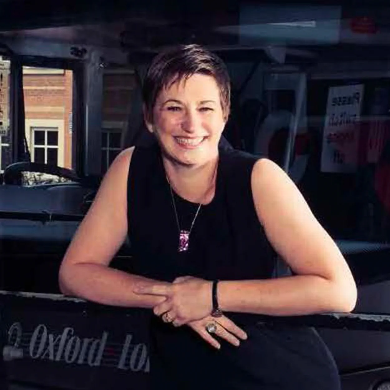 Georga Godwin is a white woman with cropped light brown hair. She is smiling and wears a short-sleeved black dress and gold necklace. She stands in front of a coach from Oxford to London Victoria.