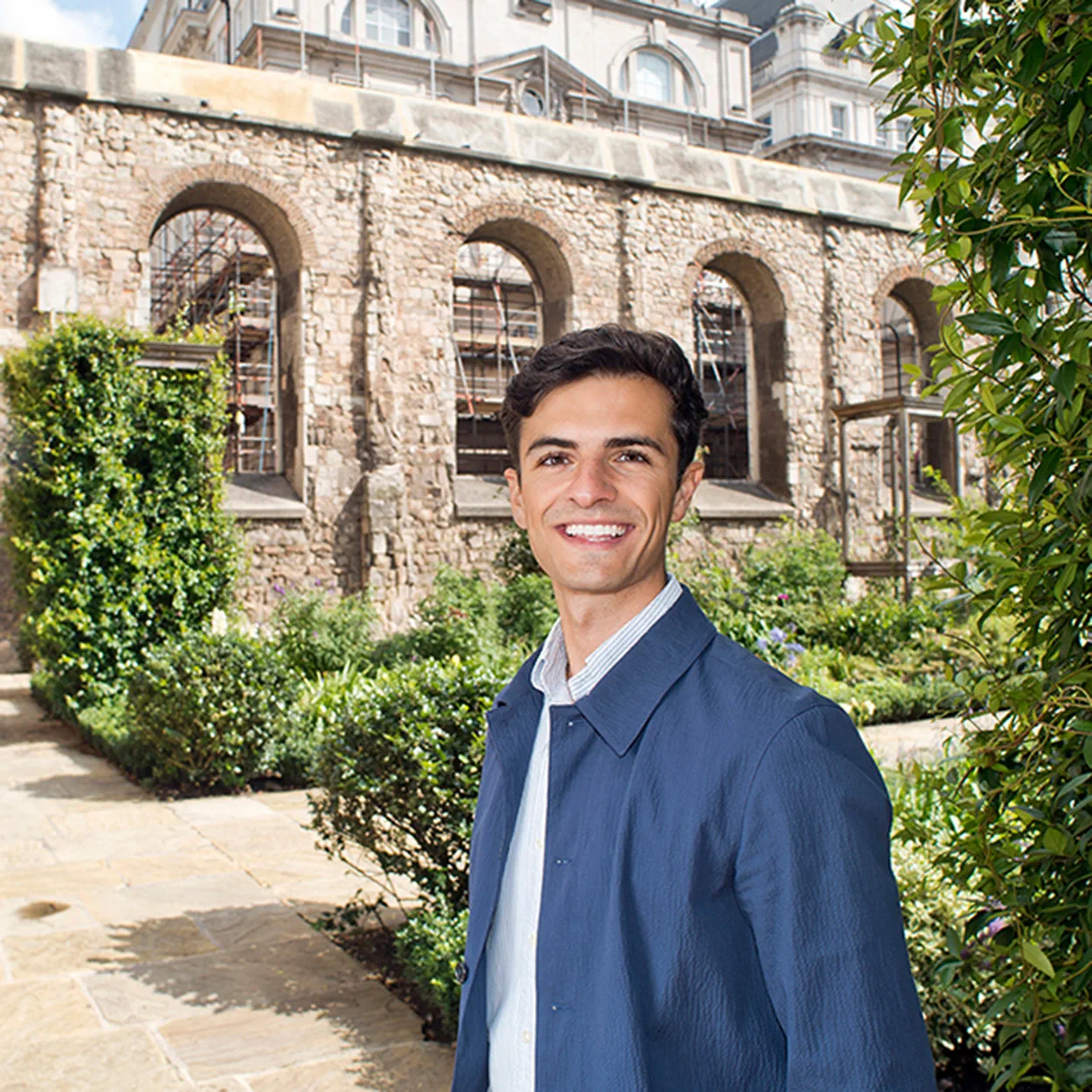 Justin Farrance is a man with short, curly dark brown hair. He is smiling widely and stands in a courtyard garden, surrounded by old stone walls. He wears an open-collar white shirt and a navy jacket.