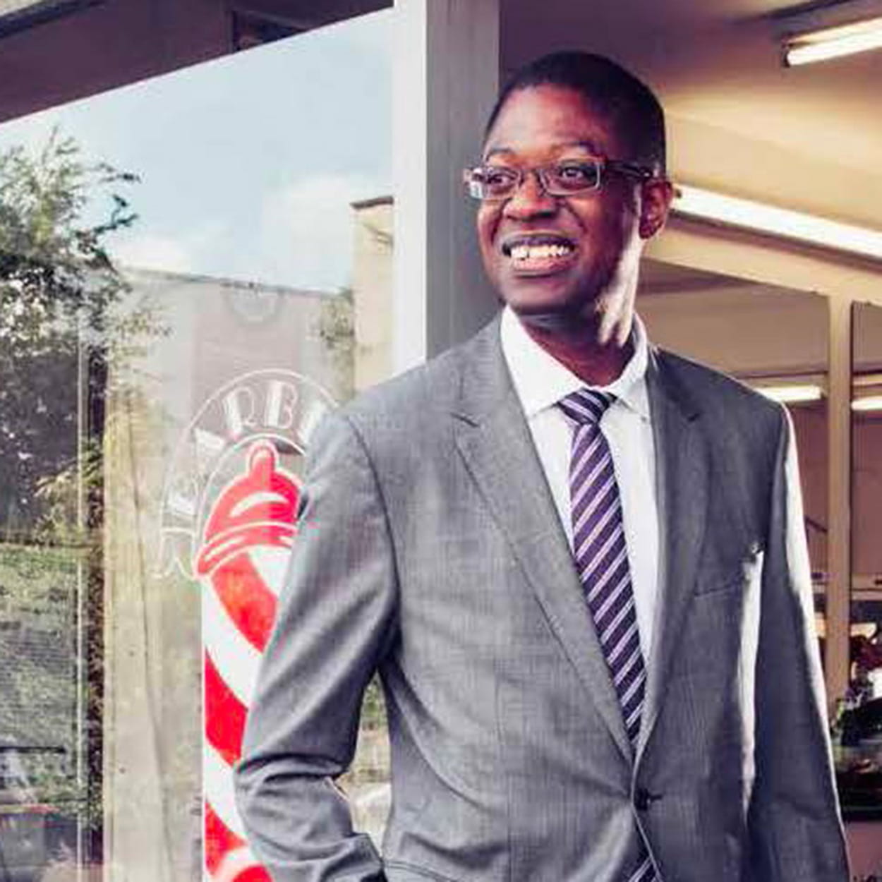 Karl Brown is a Black man with closely cropped black hair. He is smiling and wears glasses and a light grey suit, white shirt and blue and pink striped tie. He stands in the open door of a barber shop.