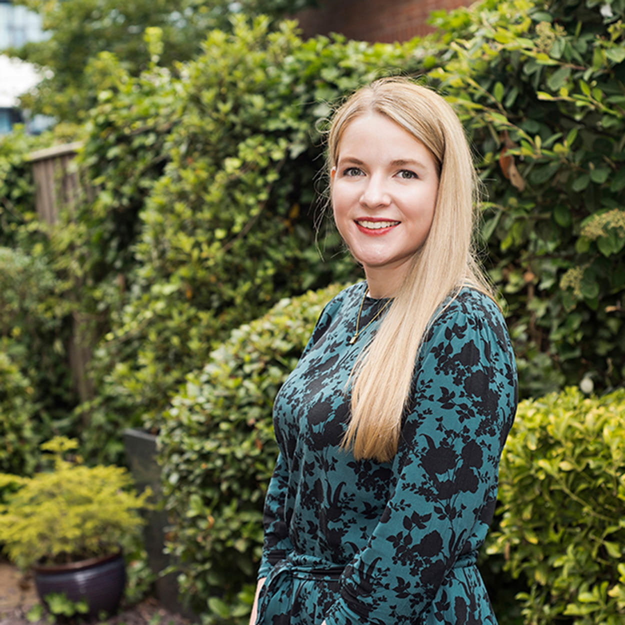 Lynette Wieland is a white woman with long, straight blonde hair. She stands outside and is smiling widely, wearing a teal and black patterned dress.