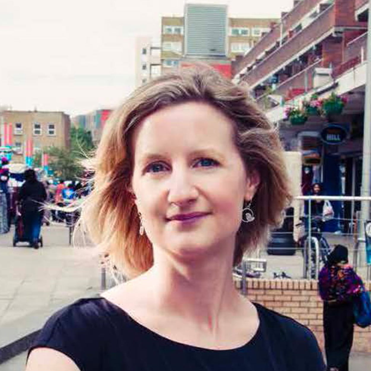 Melanie Wright is a white woman with short, straight, dark blonde hair. She is smiling and wears a black short-sleeved dress and silver earrings. She stands in front of a market place in an urban centre.