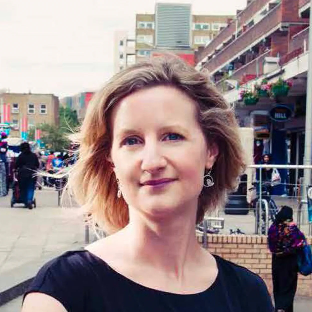 Melanie Wright is a white woman with short, straight, dark blonde hair. She is smiling and wears a black short-sleeved dress and silver earrings. She stands in front of a market place in an urban centre.