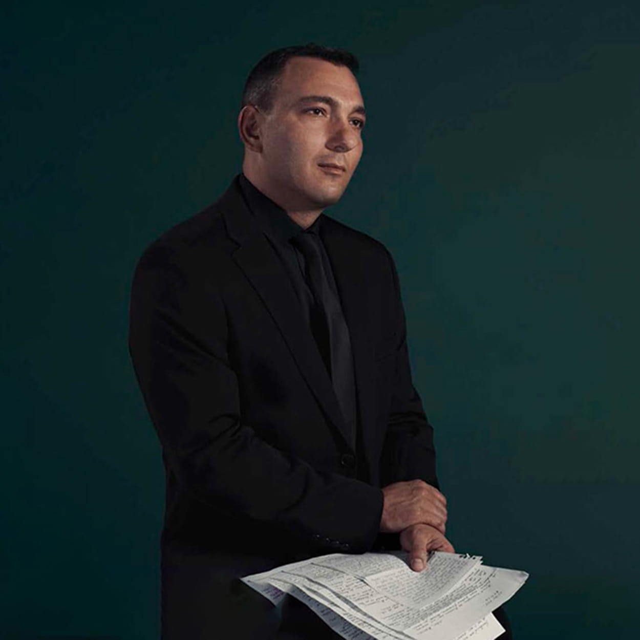 Mihail Florin Gavrilla is a white man with short black hair. He wears a black suit, black shirt and black tie, and sits holding letters from his mother.