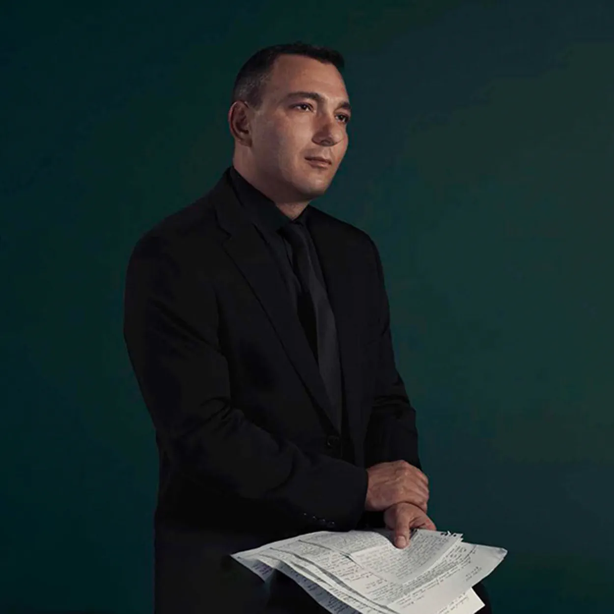 Mihail Florin Gavrilla is a white man with short black hair. He wears a black suit, black shirt and black tie, and sits holding letters from his mother.