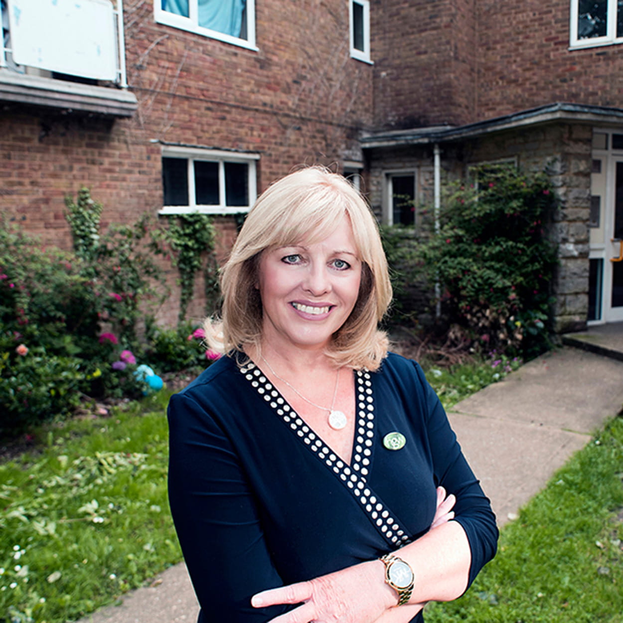 Nicola Lowe is a white woman with short, wavy blonde hair. She is smiling widely and stands in the garden outside social housing. She wears a navy wrap dress and a gold necklace and watch.