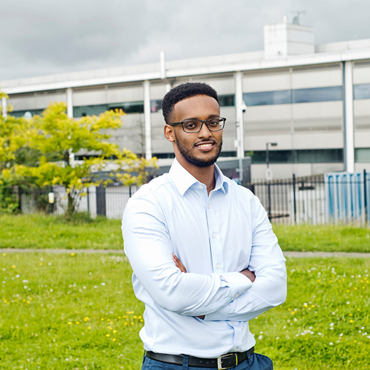 Qalid Mohamed is a Black man with short, black afro hair. He is smiling and stands in a park outside of an industrial building. He wears a pale blue shirt and glasses.