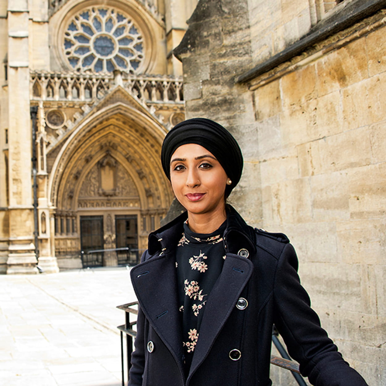 Salma Maqsood is a woman of South Asian heritage who wears a black headscarf. She is smiling and stands in front of a cathedral. She wears a black coat over a black floral dress.