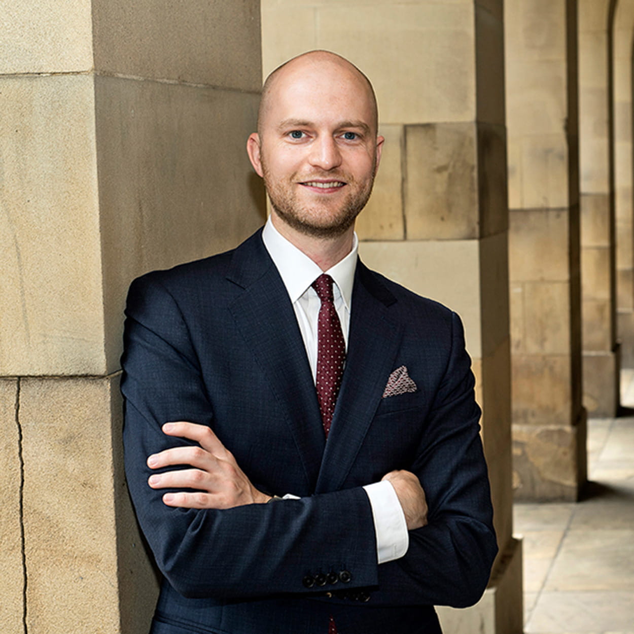 Sam Butler is a white man with closely shaved hair and a short beard. He is smiling and wears a black suit, white shirt and dark red tie.