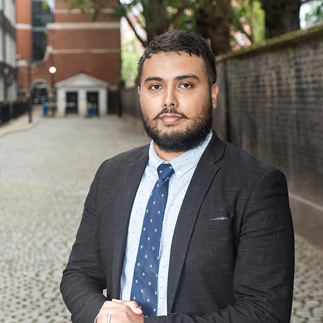 Shaheen Mamun is a man of South Asian heritage with short, dark brown hair and a medium-length beard. He stands on a cobbled street with his hands clasped. He wears a dark grey suit, white shirt and navy patterned tie.