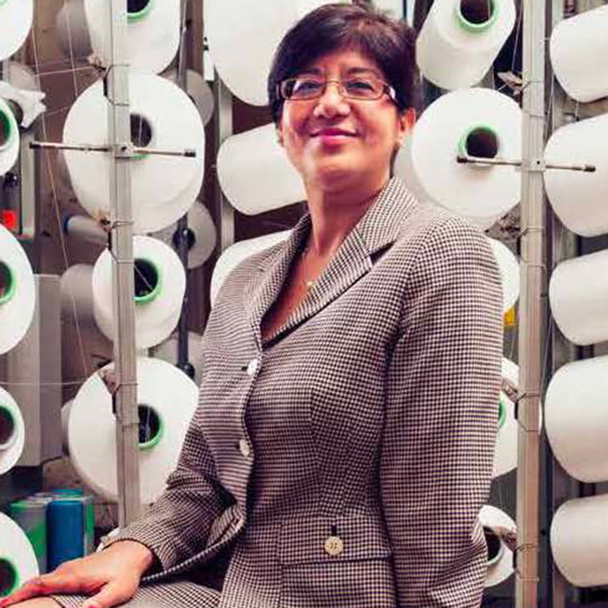 Shama Gupta is a woman of South Asian heritage with short, straight, dark brown hair. She is smiling and wears glasses and a light brown tweed suit.