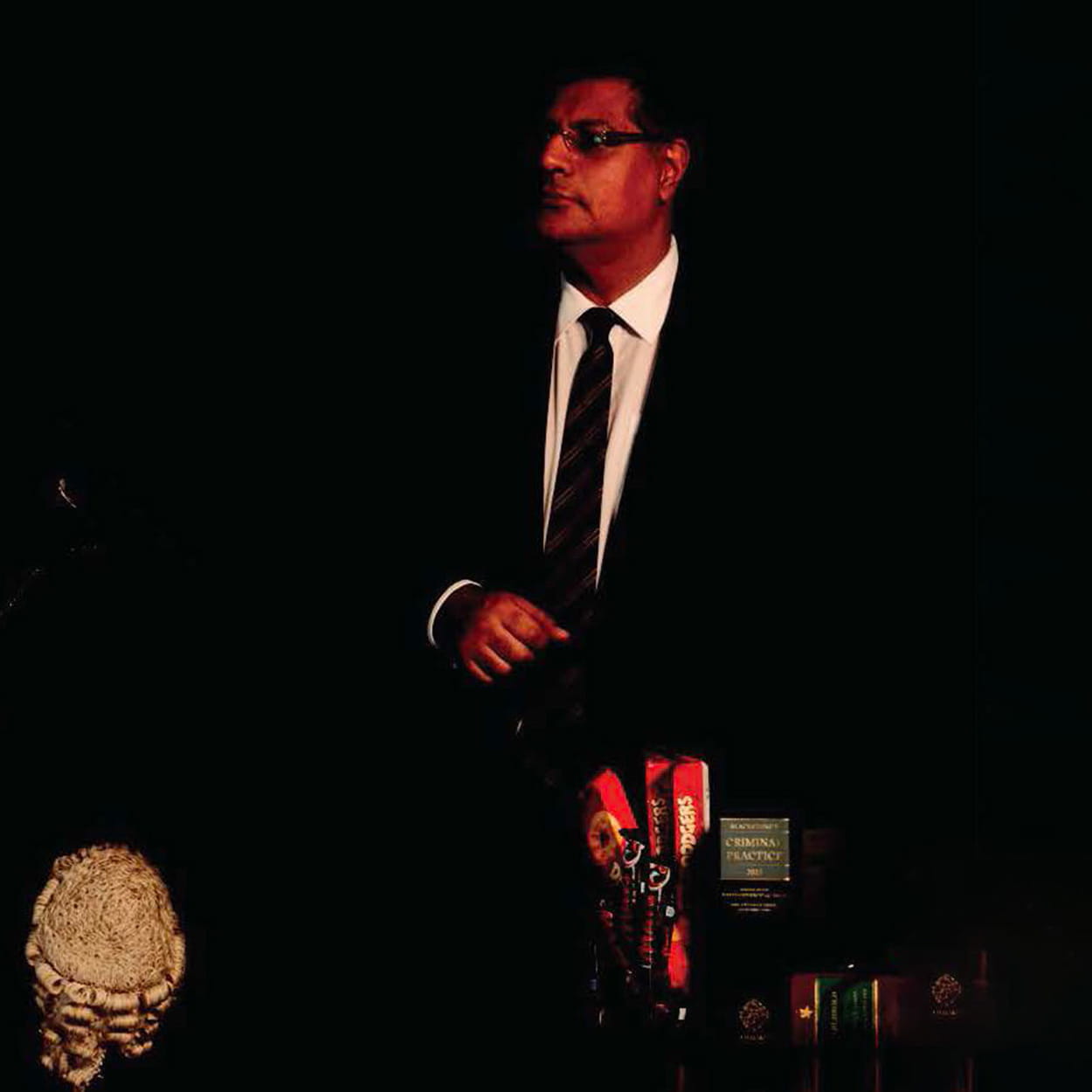 Tan Ikram is a man of South Asian heritage with short, black hair. He wears glasses and a black suit, white shirt and black striped tie. He stands behind a judge's wig and several accolades.