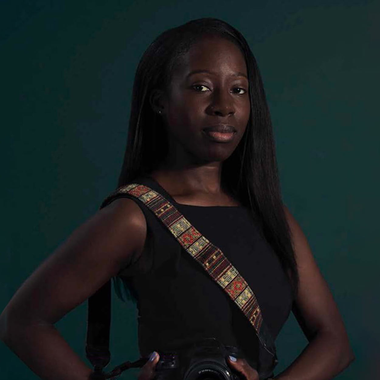 Whitney Joseph is a Black woman with long, straight black hair. She wears a black sleeveless dress and holds a digital camera.