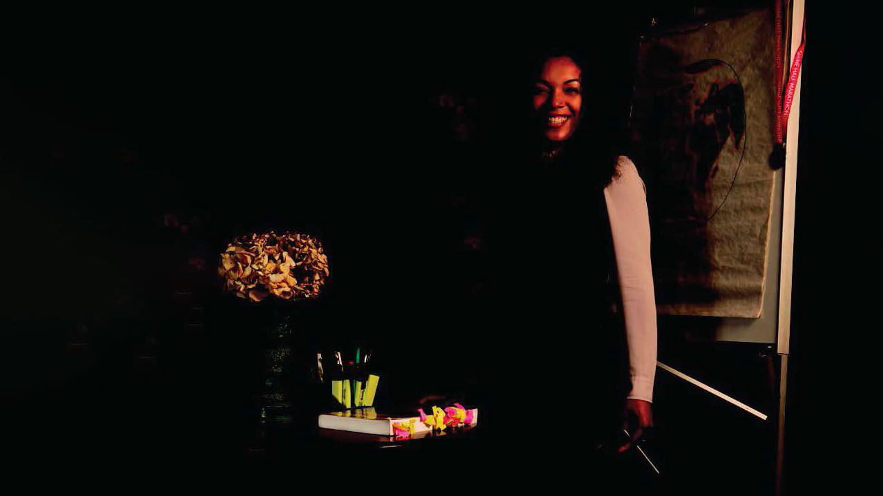 Adele Edwin-Lamerton is a Black woman with long, dark, wavy hair. She is smiling and wears a long-sleeved white blouse and black dress. She stands holding a pen in front of a desk with a heavily tabbed textbook.