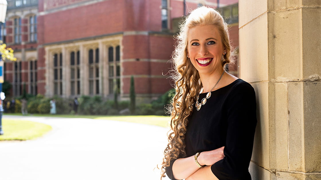Alice Kinder is a white woman with long, curly blonde hair. She is smiling widely and stands outside, in front of a grand, brick building. She wears a long-sleeved black dress and a silver necklace.