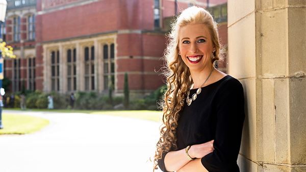 Alice Kinder is a white woman with long, curly blonde hair. She is smiling widely and stands outside, in front of a grand, brick building. She wears a long-sleeved black dress and a silver necklace.