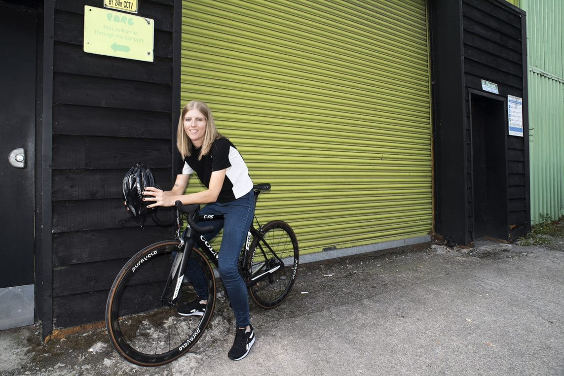 Clare is a blonde white woman perched on a black bicycle wearing a sporty t-shirt and skinny jeans with black and white trainers. She is holding a black bicycle helmet, and sits in front of a chartreuse shutter door.