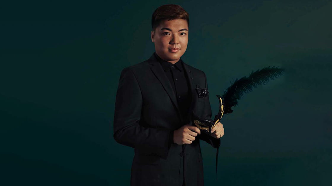 Billy Yu Lok Ng is a man of East Asian descent with short, dark brown hair. He wears a black suit with a black shirt and holds a gold and black feathered theatre mask.