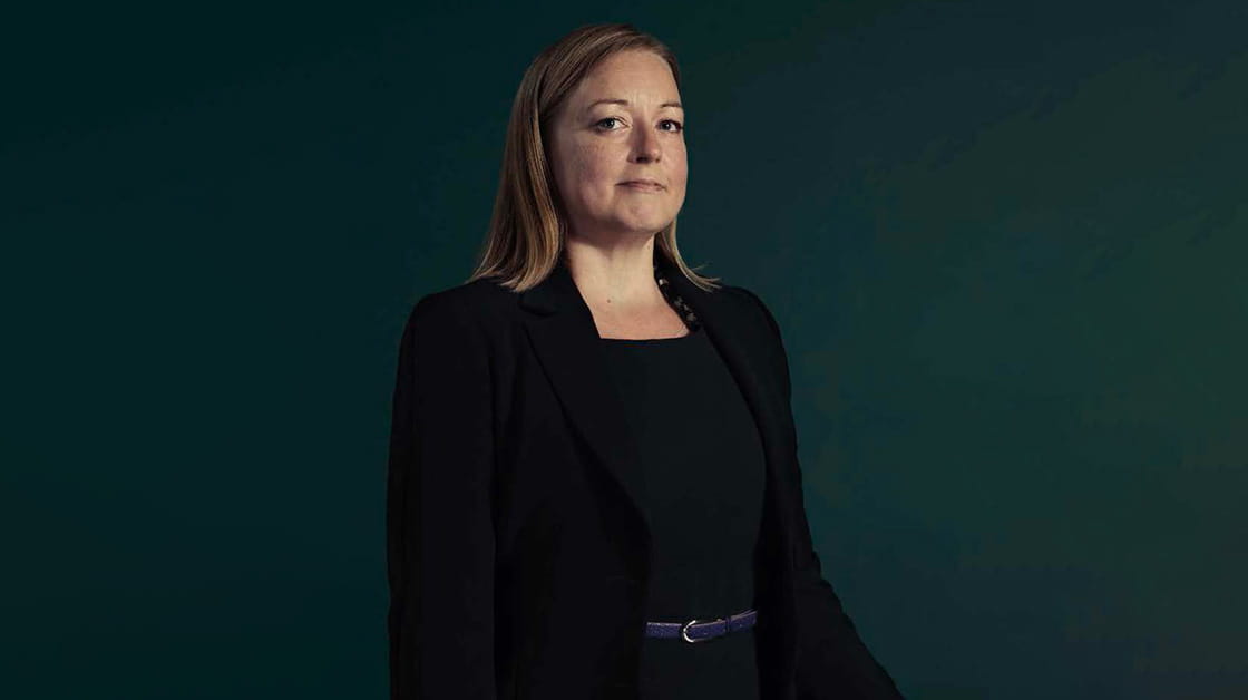 Gemma Lodge was a white woman with long, straight, dark blonde hair. She wears a black dress with a black blazer and leather belt.