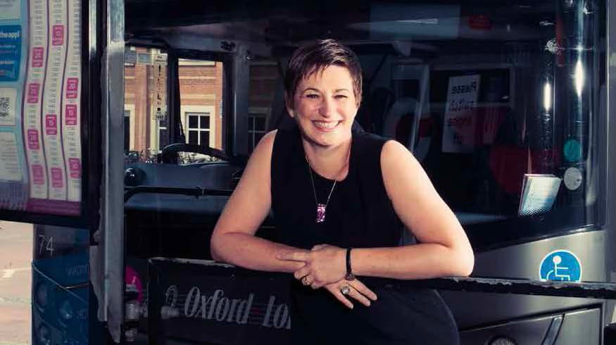 Georga Godwin is a white woman with cropped light brown hair. She is smiling and wears a short-sleeved black dress and gold necklace. She stands in front of a coach from Oxford to London Victoria.