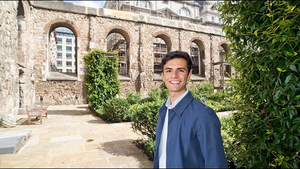Justin Farrance is a man with short, curly dark brown hair. He is smiling widely and stands in a courtyard garden, surrounded by old stone walls. He wears an open-collar white shirt and a navy jacket.