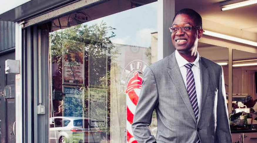 Karl Brown is a Black man with closely cropped black hair. He is smiling and wears glasses and a light grey suit, white shirt and blue and pink striped tie. He stands in the open door of a barber shop.