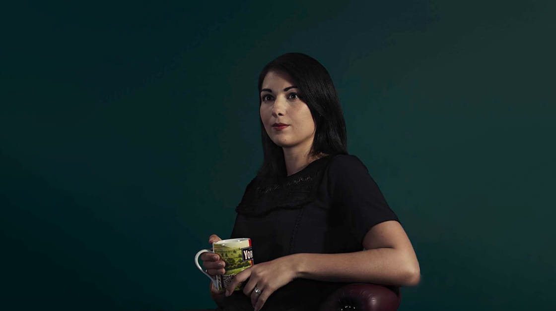 Kira Wilkinson is a white woman with medium-length, straight dark brown hair. She wears a black dress and holds a mug of Yorkshire Tea.