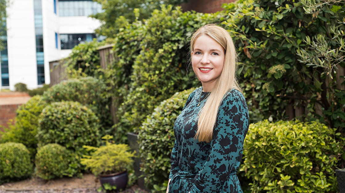 Lynette Wieland is a white woman with long, straight blonde hair. She stands outside and is smiling widely, wearing a teal and black patterned dress.