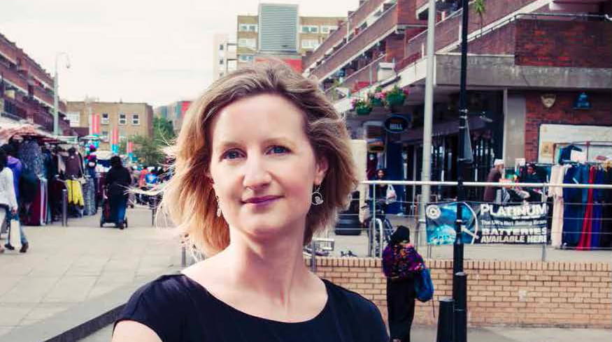 Melanie Wright is a white woman with short, straight, dark blonde hair. She is smiling and wears a black short-sleeved dress and silver earrings. She stands in front of a market place in an urban centre.
