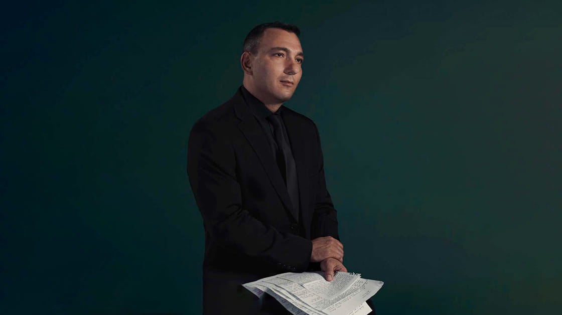Mihail Florin Gavrilla is a white man with short black hair. He wears a black suit, black shirt and black tie, and sits holding letters from his mother.