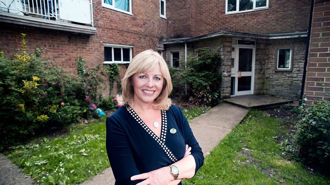 Nicola Lowe is a white woman with short, wavy blonde hair. She is smiling widely and stands in the garden outside social housing. She wears a navy wrap dress and a gold necklace and watch.
