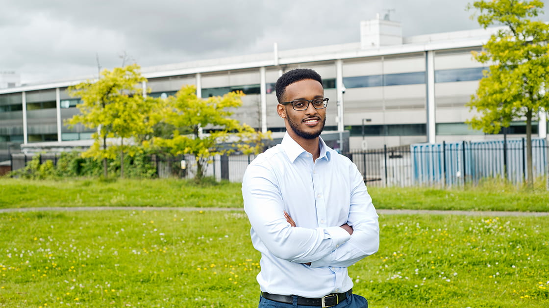 Qalid Mohamed is a Black man with short, black afro hair. He is smiling and stands in a park outside of an industrial building. He wears a pale blue shirt and glasses.