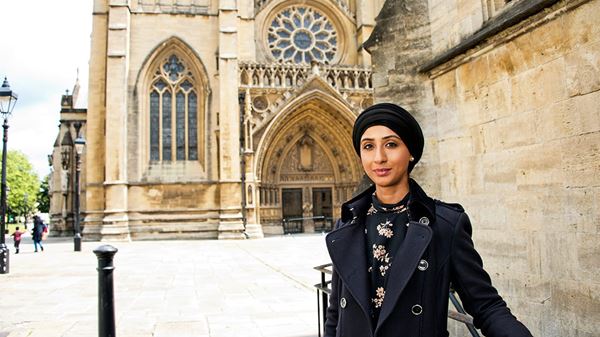 Salma Maqsood is a woman of South Asian heritage who wears a black headscarf. She is smiling and stands in front of a cathedral. She wears a black coat over a black floral dress.