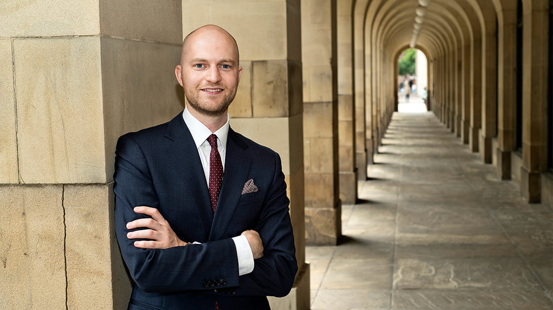 Sam Butler is a white man with closely shaved hair and a short beard. He is smiling and wears a black suit, white shirt and dark red tie.