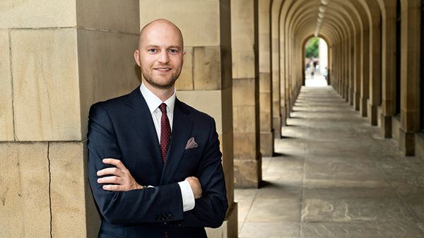Sam Butler is a white man with closely shaved hair and a short beard. He is smiling and wears a black suit, white shirt and dark red tie.