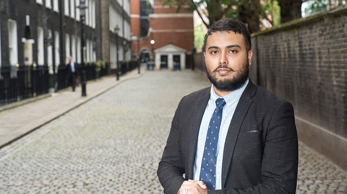 Shaheen Mamun is a man of South Asian heritage with short, dark brown hair and a medium-length beard. He stands on a cobbled street with his hands clasped. He wears a dark grey suit, white shirt and navy patterned tie.