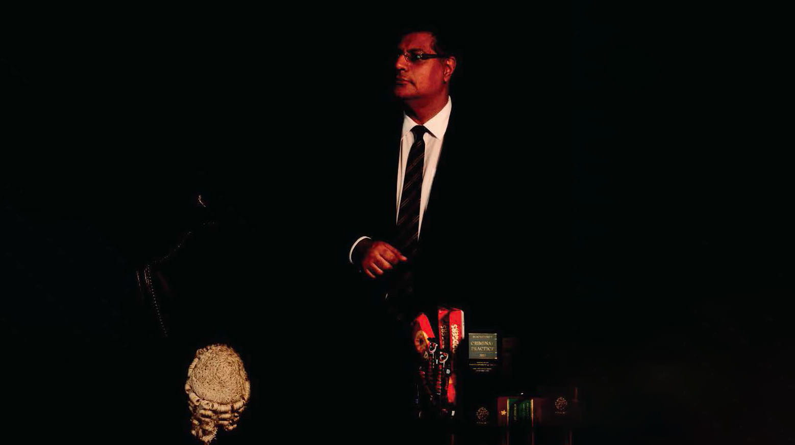 Tan Ikram is a man of South Asian heritage with short, black hair. He wears glasses and a black suit, white shirt and black striped tie. He stands behind a judge's wig and several accolades.