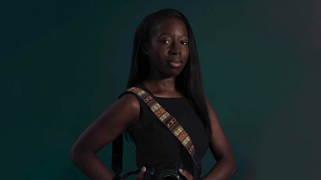 Whitney Joseph is a Black woman with long, straight black hair. She wears a black sleeveless dress and holds a digital camera.