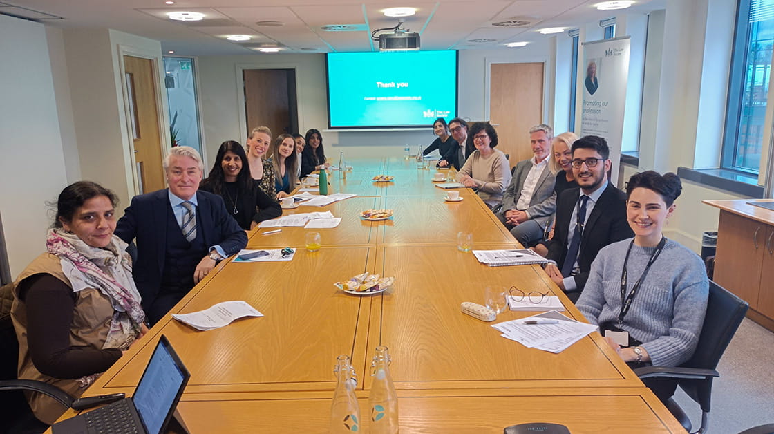 Law Society president, Lubna Shuja, and other members at roundtable in Birmingham to discuss socio-economic diversity in the legal profession. Thirteen people sitting around boardroom table, smiling at the camera