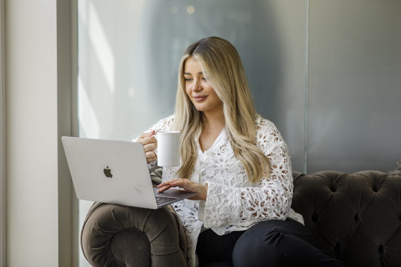 A young professional sits on a sofa browsing a laptop and holding a mug of tea.