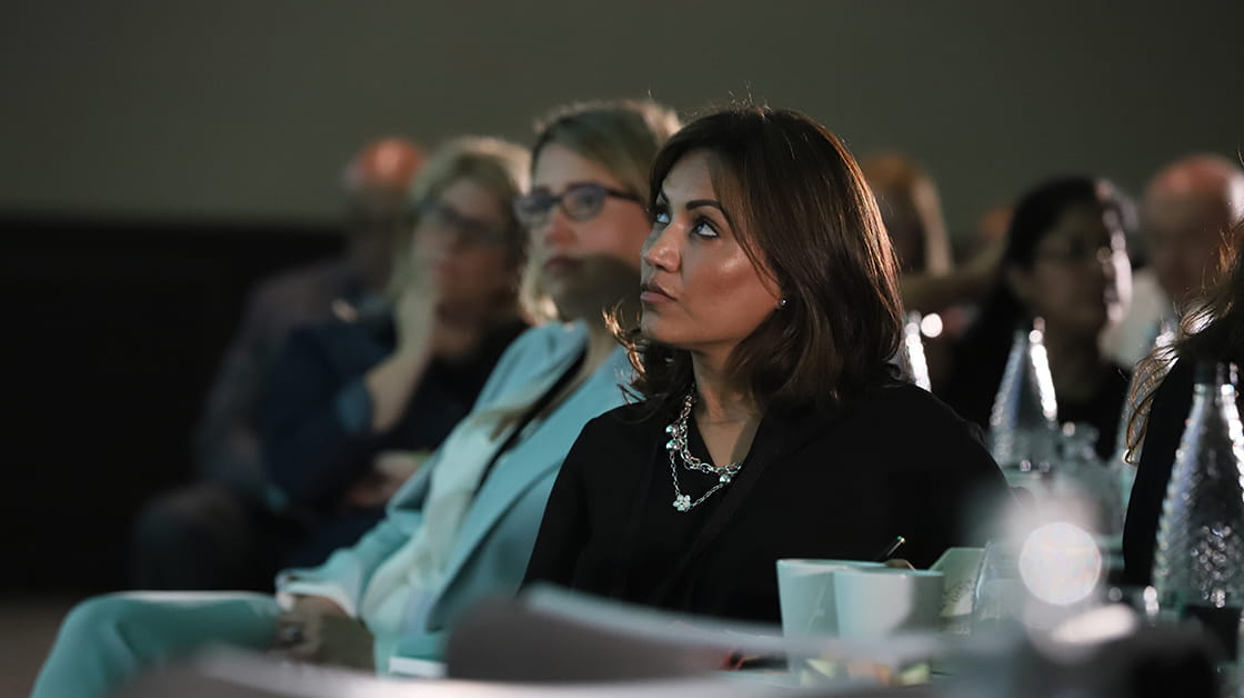 A woman sits watching presentation in darkened conference room. She's wearing a silver chain with flower detail over a dark suit. Behind her sit other attendees.