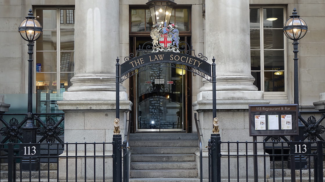 The Law Society entrance at 113 Chancery Lane, London. Stone building with pillars and iron railings outside. Under an arch with sign bearing the Law Society crest, steps lead up to glass doors with lights inside.