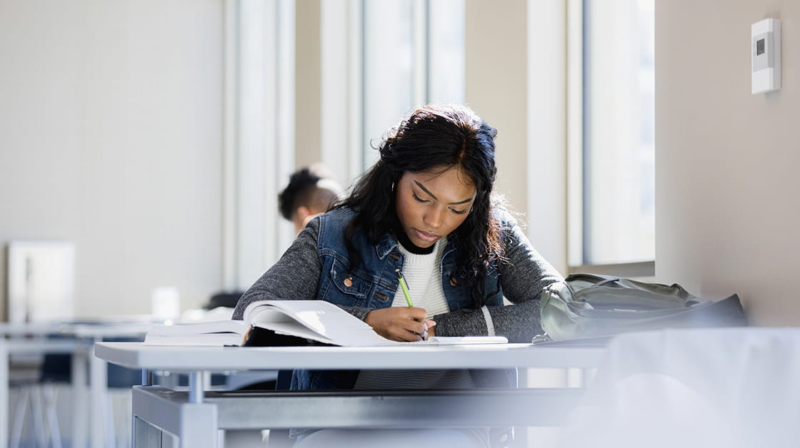 A black woman is studying at a desk, crouching over a book.