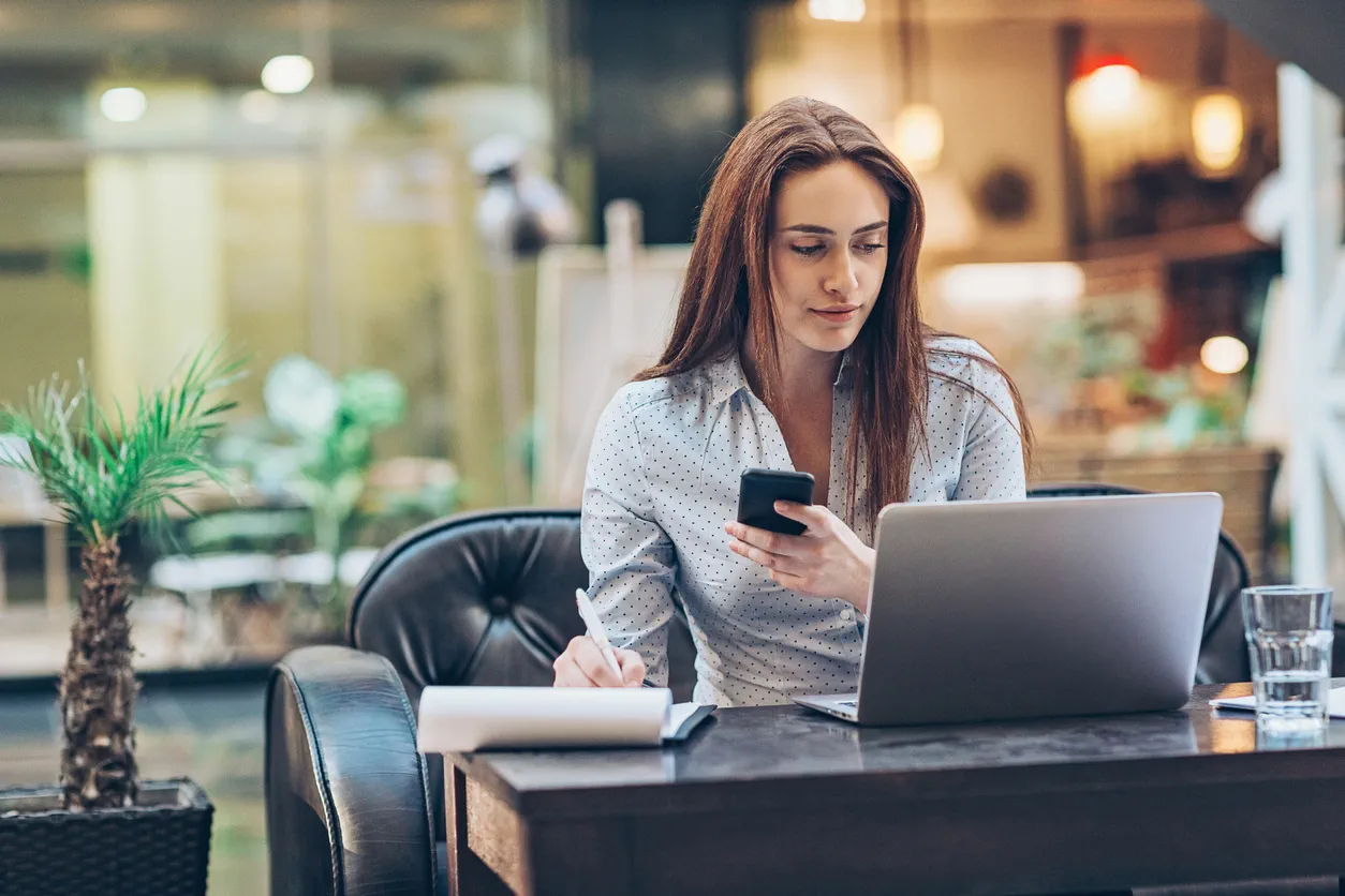 A woman sits at a desk with a closed mouth smile. She is looking at a laptop, holding a phone and writing on a piece of paper.