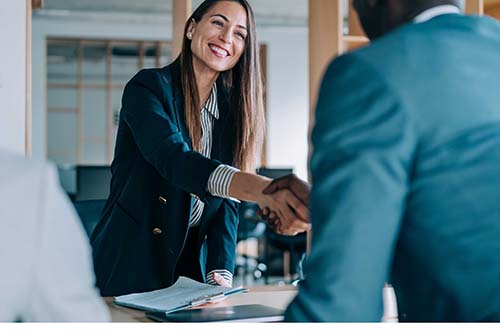 Professional woman shakes hands with employee across desk (she is holding a clipboard and has long, straight hair)