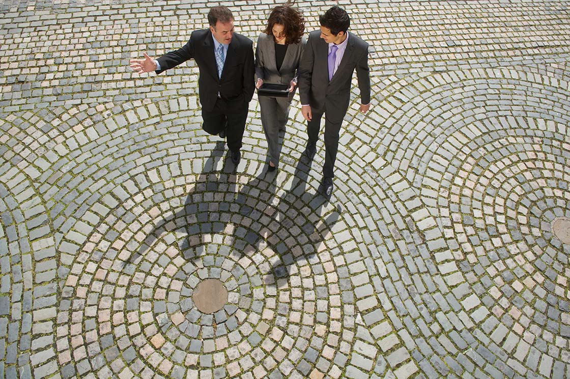 Bird's eye view of three professionals walking outside, they cast a strong shadow on the ground, which is paved in swirling patterns